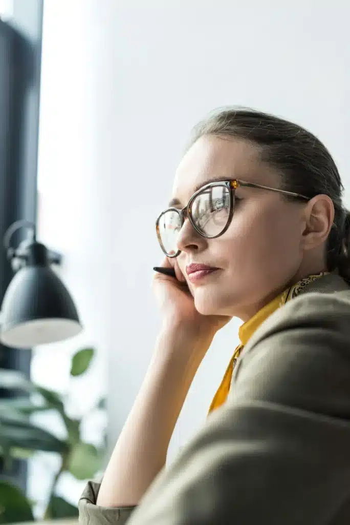 beautiful pensive businesswoman in eyeglasses looking away at workplace