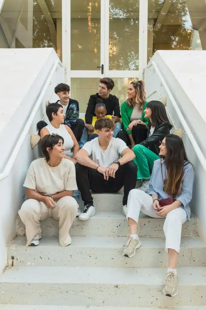 Group of happy diverse friends sitting on stairs of glass door entrance in daylight