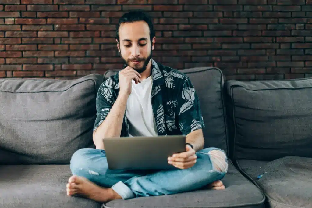 man in casual wear sitting on couch barefoot and reading laptop