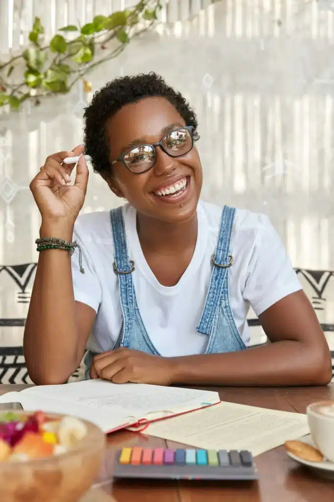 Photo of cheerful black woman with broad smile, ponders on idea for solution, plans costs and writes