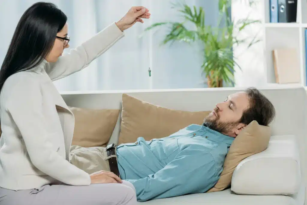 side view of young hypnotist with pendulum and bearded man with closed eyes lying in couch during