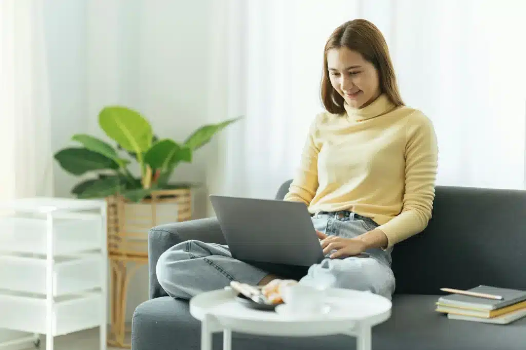 Smiling beautiful young asian ethnicity woman sitting on sofa with computer on lap, thinking of dail