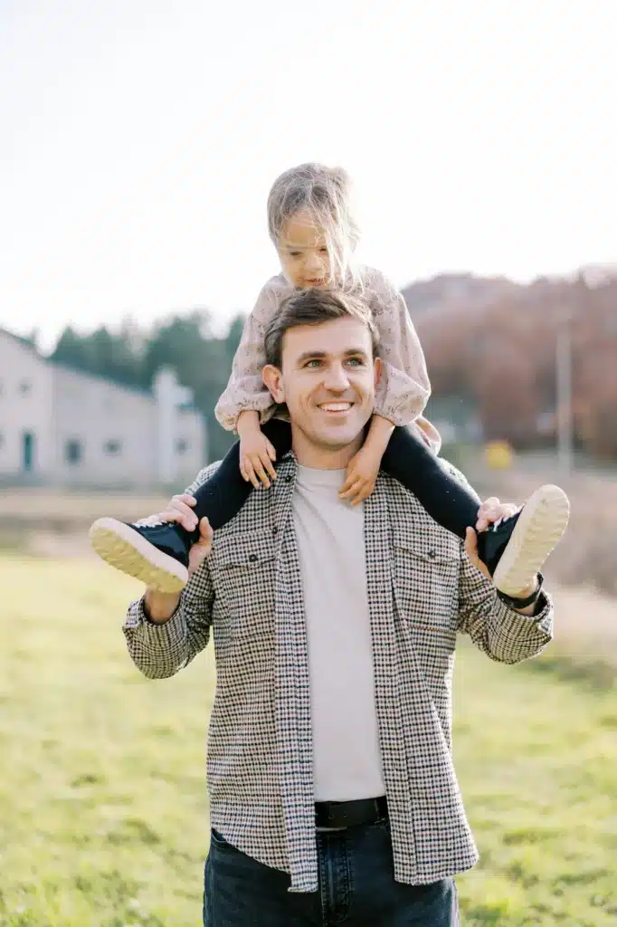 Smiling dad carries little girl on his shoulders across the meadow, looking to the side