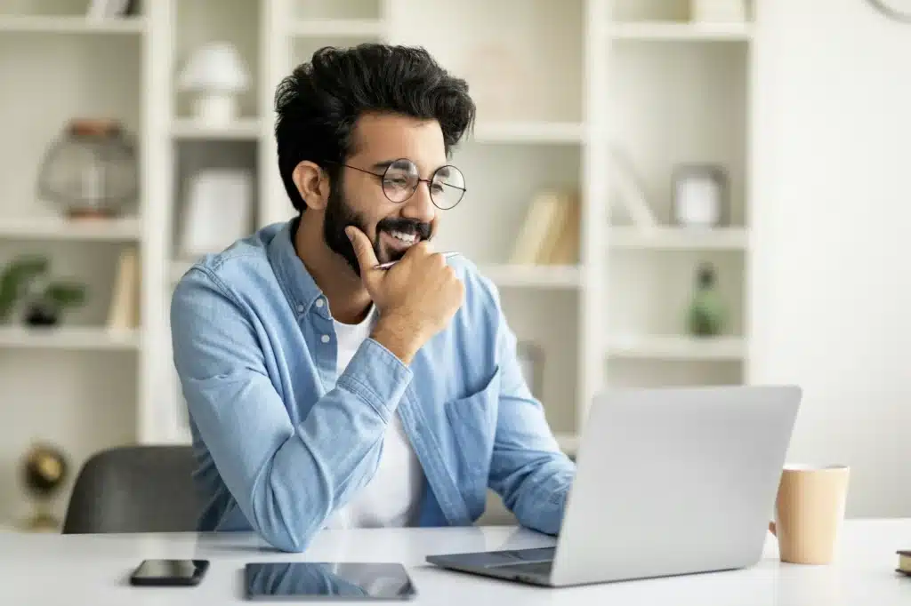 Telecommuting Concept. Smiling Indian Man Working Online With Laptop At Home Office