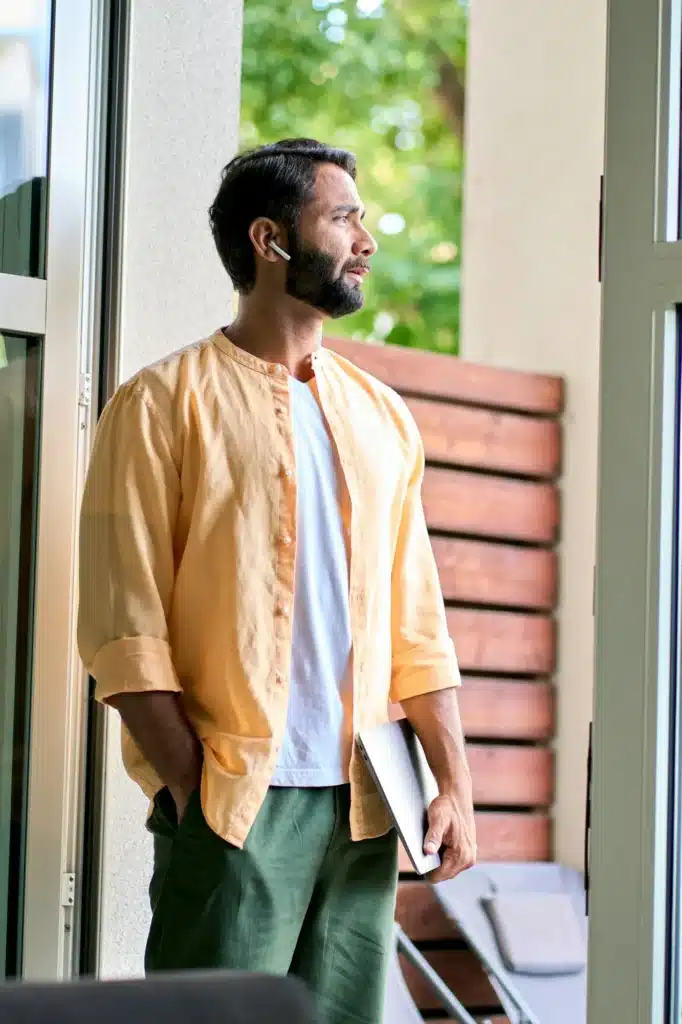 Thoughtful indian man holding laptop standing at doorway and thinking.