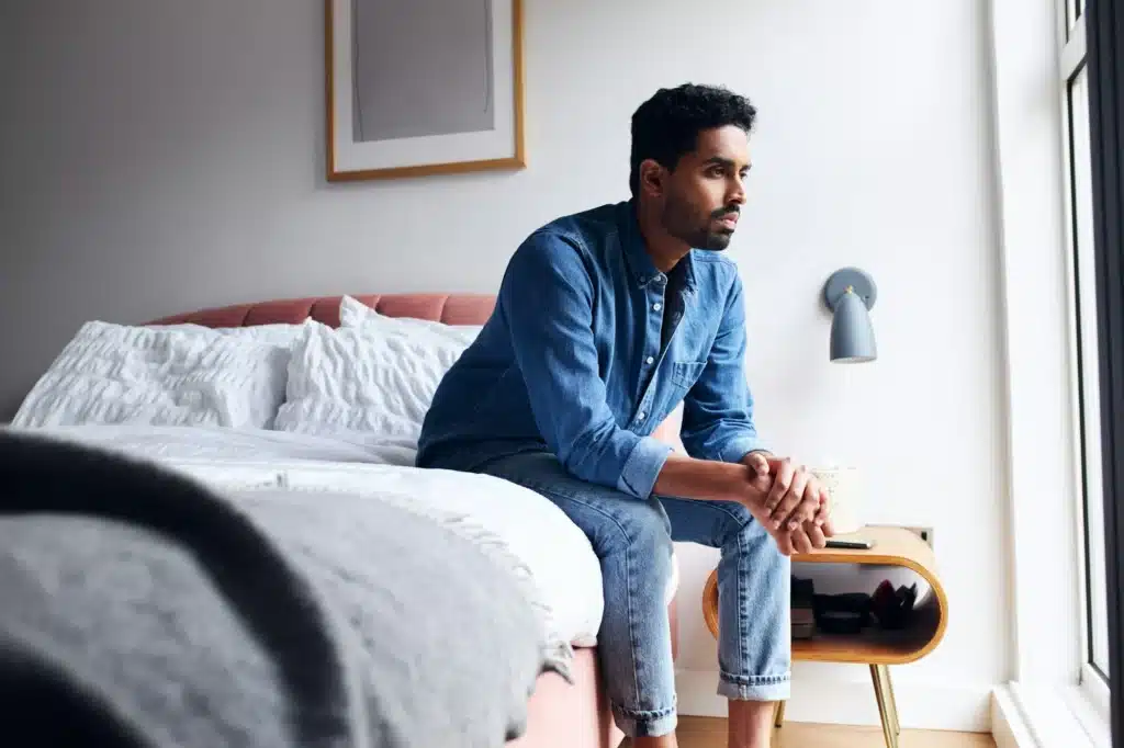 Unhappy young man with mental health issues sitting on edge of bed and staring out of window