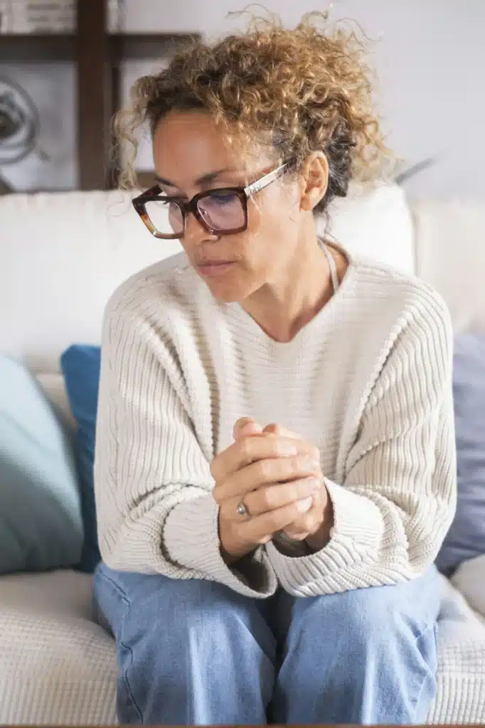 Upset and sad adult woman sitting on sofa with clasped hands looking down. Anxiety and stress concep