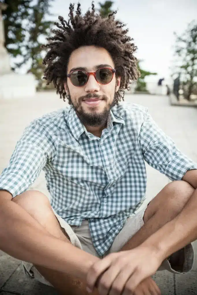 Young African man sitting on the ground with hair braids and sunglasses wearing a shirt and shorts