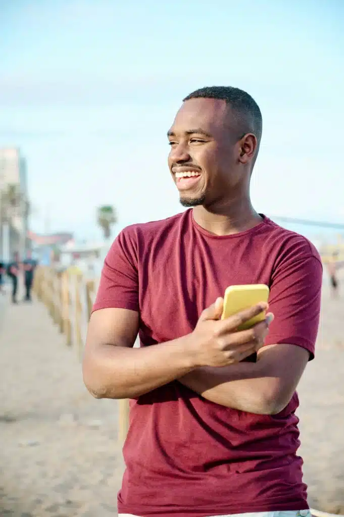 young black man at the beach smiling and using his smartphone