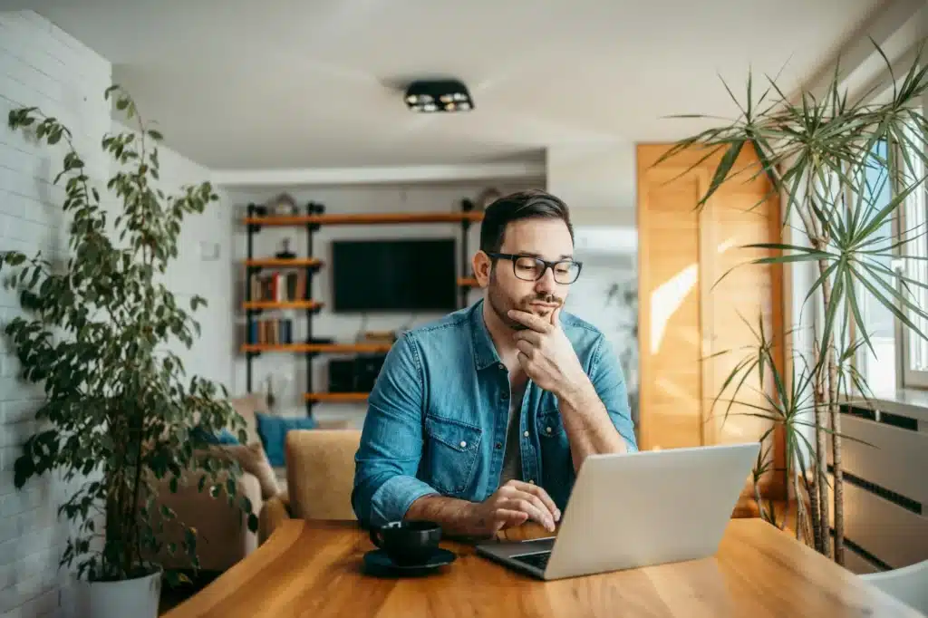 Portrait of a casual businessman thinking while looking at laptop screen, keeping hand on chin.