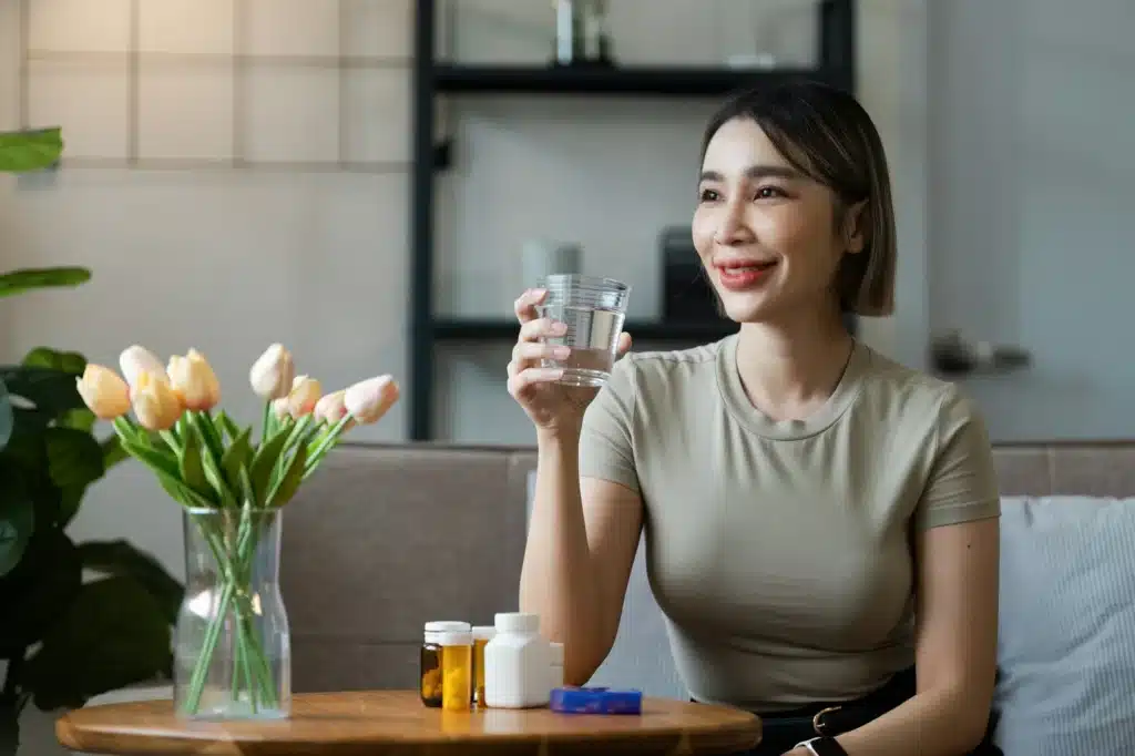 Woman Sitting on Sofa Holding a Glass of Water with Medication Bottles on Table in Cozy Living Room