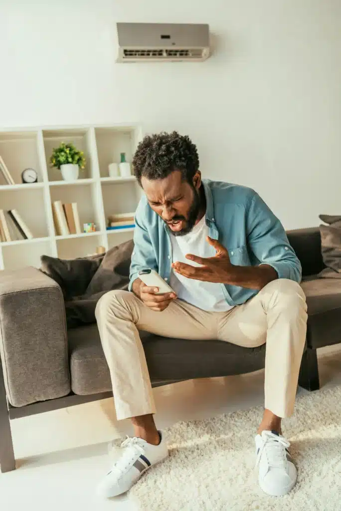 irritated african american man holding air conditioner remote controller while suffering from heat