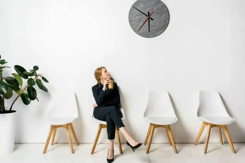 young businesswoman holding coffee to go and looking at clock while waiting in line